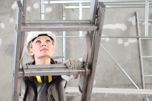 woman construction worker builder on ladder wearing white helmet and hearing protection headphones on interior site building background